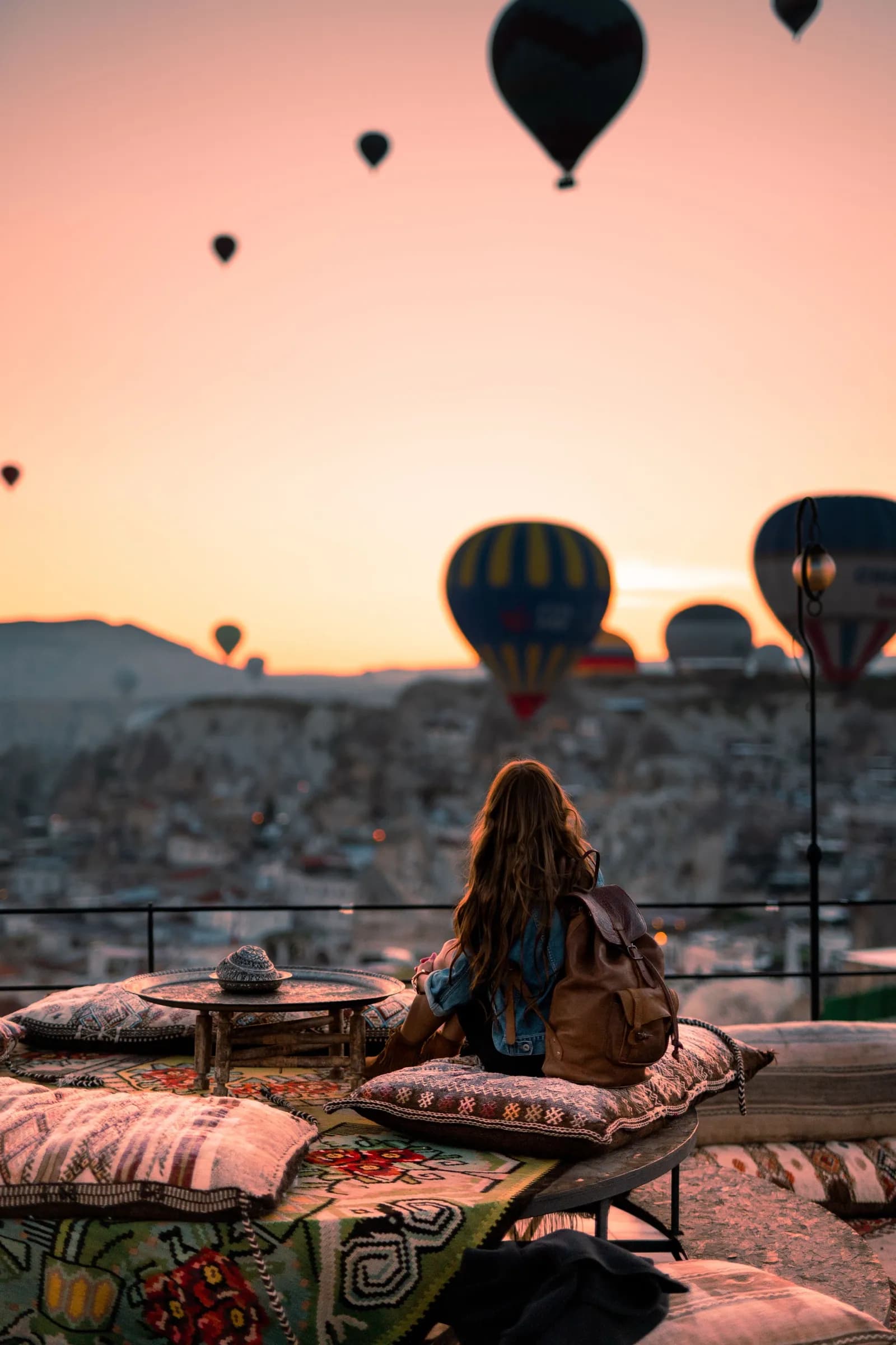 A woman on a cave-hotel terrace watching hot-air balloons rise at sunrise